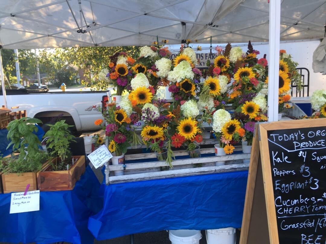 117326750_742791672958520_9188695172725500452_n Colorful flower display at a farmer's market featuring sunflowers, zinnias, and hydrangeas, with a sign listing available organic produce like kale, peppers, and eggplant. A wooden box with herbs is also visible on a blue tablecloth under a tent.