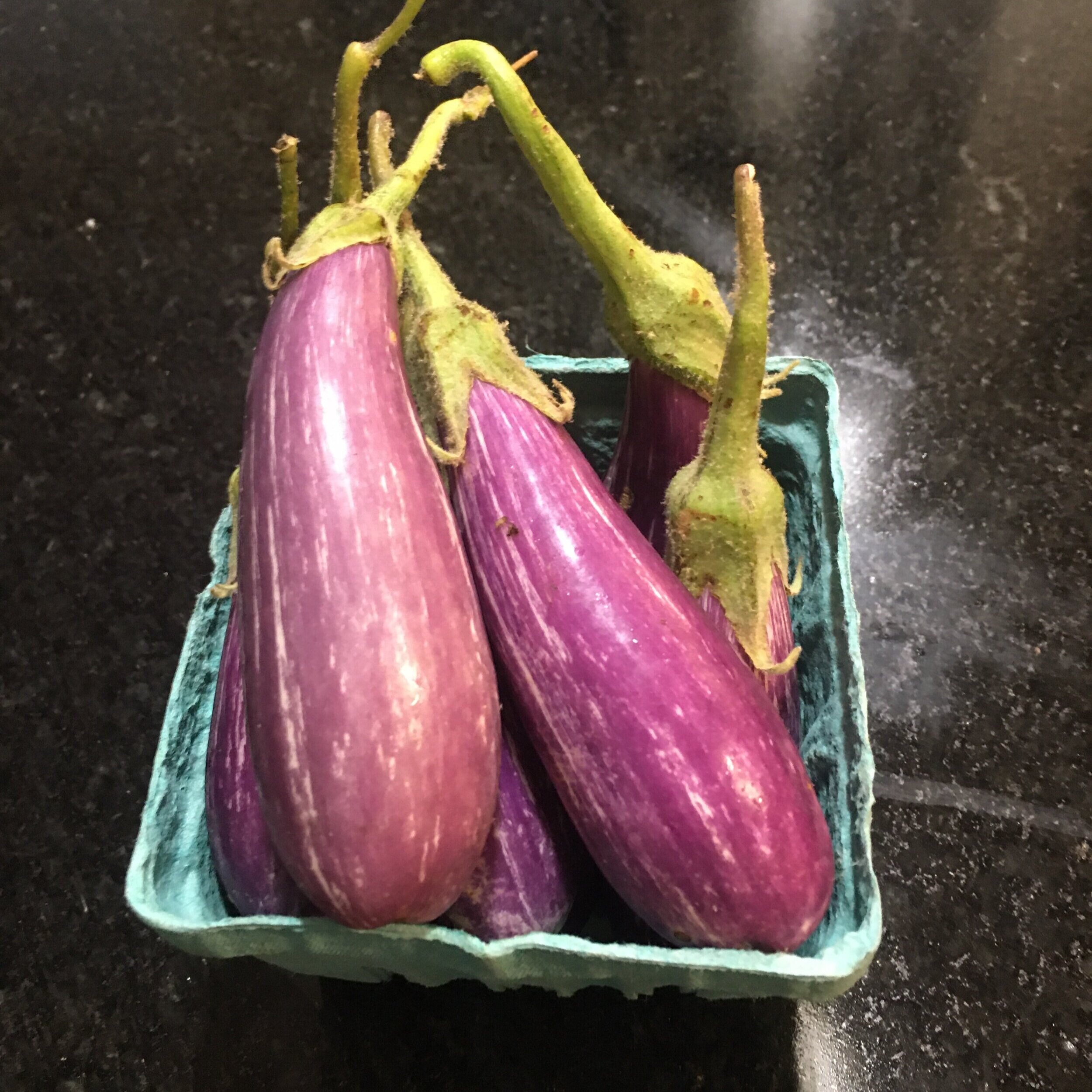 Fresh purple eggplants displayed in a green cardboard container on a dark countertop, showcasing their glossy skin and vibrant color, ideal for healthy recipes and culinary dishes.