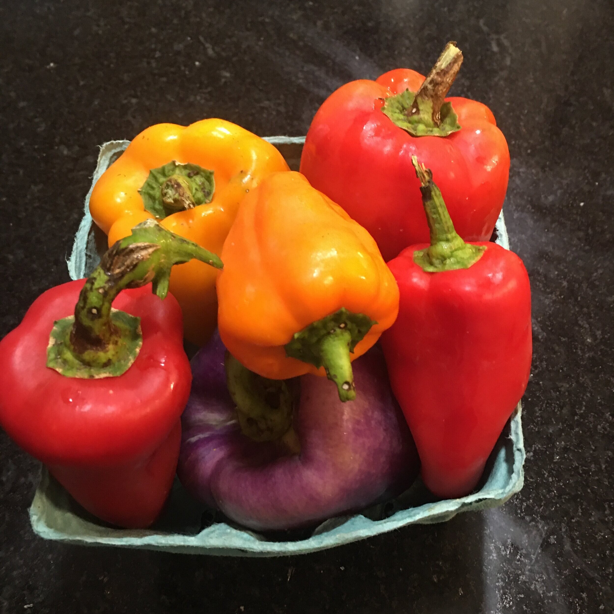 Colorful assortment of bell peppers including red, yellow, and purple varieties, arranged in a green cardboard container on a dark countertop. Perfect for healthy recipes and vibrant dishes.